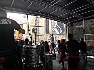 Hudebníci na venkovním pódiu s bicími, vlajkami a budovami v pozadí.