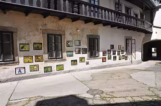 Obrazy vystavené na zdi starého kamenného domu s dřevěným balkonem na dlážděném nádvoří