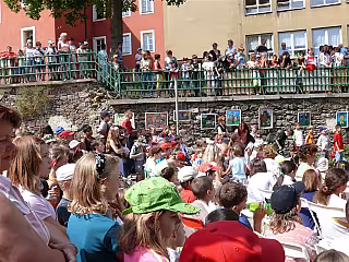 Skupiny dětí a dospělých na nádvoří před červenou budovou během venkovní akce.