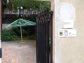 Otevřené černé dveře vedoucí do zahrady se zeleným slunečníkem a terakótovou dlažbou.