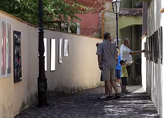 Lidé prohlížejí venkovní výstavu fotografií na zdi v úzké dlážděné uličce.