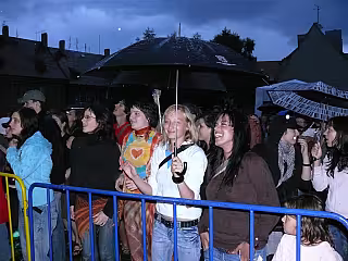 Večerní dav lidí u modré zábradlí na venkovním festivalu nebo eventos.