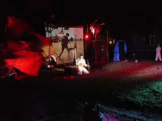 Noční venkovní divadelní představení s herci, červeným a zeleným světlem a kulisami.