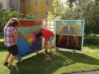 Dvě osoby malují velké barevné obrazy na trávníku před žlutou budovou.