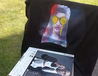 Černé tričko s potiskem obličeje a časopis na trávě venku.