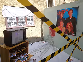 Pokoj s televizorem, žlutou výstražnou páskou a fotografií čtyř lidí na zdi.
