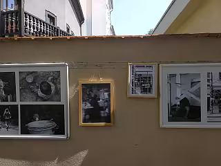 Venkovní výstava černobílých fotografií na béžové zdi budovy.