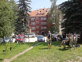 Lidé sedí na trávníku před červenou cihlovou budovou, vedle zaparkovaných aut a stromů.