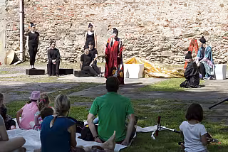 Středověké divadelní představení na hradě, herci v kostýmech, diváci sedí na trávě.