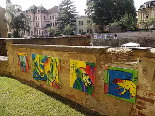 Kamenná zeď s barevnými graffiti malbami zvířat, za ní historické budovy a stromy.