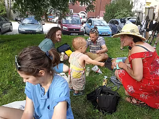Skupina lidí sedí na trávě v parku, malé dítě stojí mezi nimi za slunečného dne.