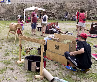 Lidé na středověkém festivalu u dřevěných konstrukcí a exponátů na trávníku.