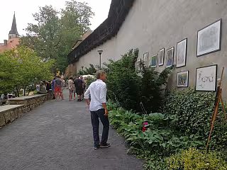 Muž prochází venkovní výstavou obrazů podél kamenné zdi obklopené zelení.