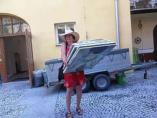 Žena v klobouku a červené sukni drží velký obraz u přívěsu na dlážděném dvorku.
