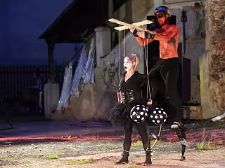 Loutkoherec v červeném ovládá marionetu na pódiu před budovou v noci.