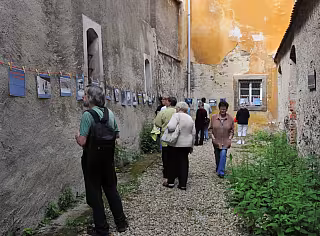Skupina lidí prochází úzkou uličkou mezi starými omítnutými zdmi budov.