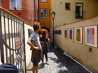 Dva lidé prohlížejí barevné obrazy vystavené na zdi úzké historické uličky.