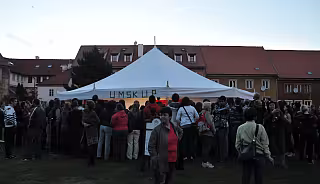 Dav lidí shromážděný kolem bílého párty stanu na trávníku před cihlovými budovami za soumraku